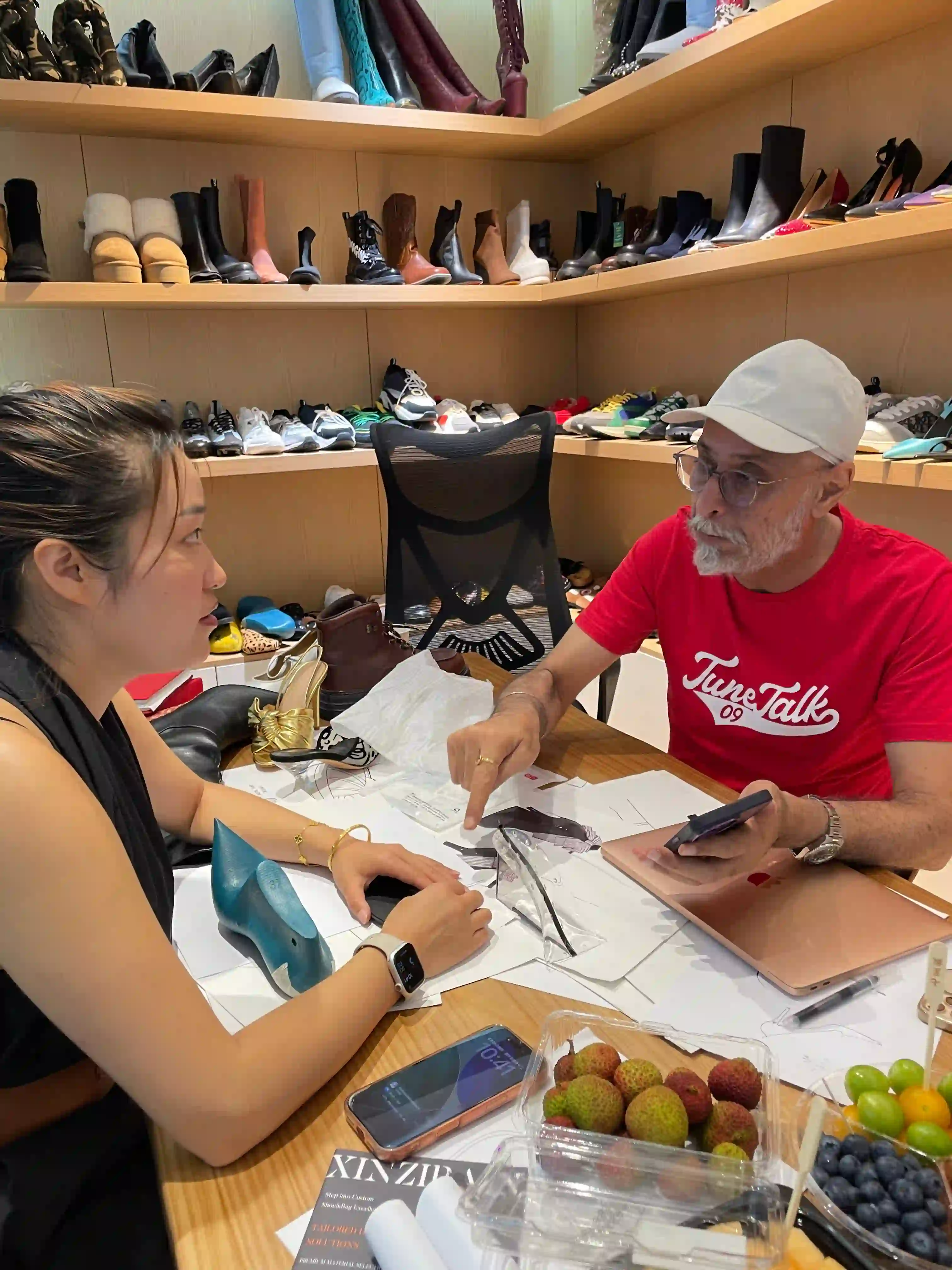 Client pointing at a design sheet on the table while a designer listens, blue-green last, black sandals and samples beside them