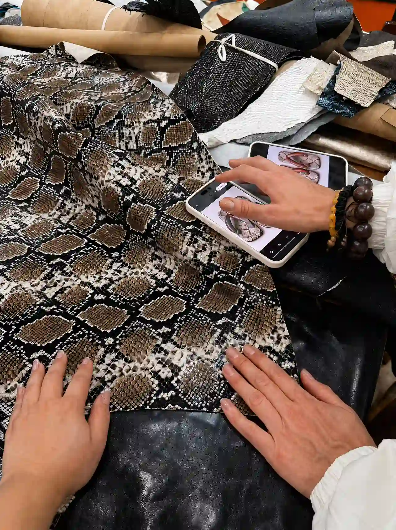 Hands spreading a large python-print leather hide while a phone on the table shows the shoe design for comparison