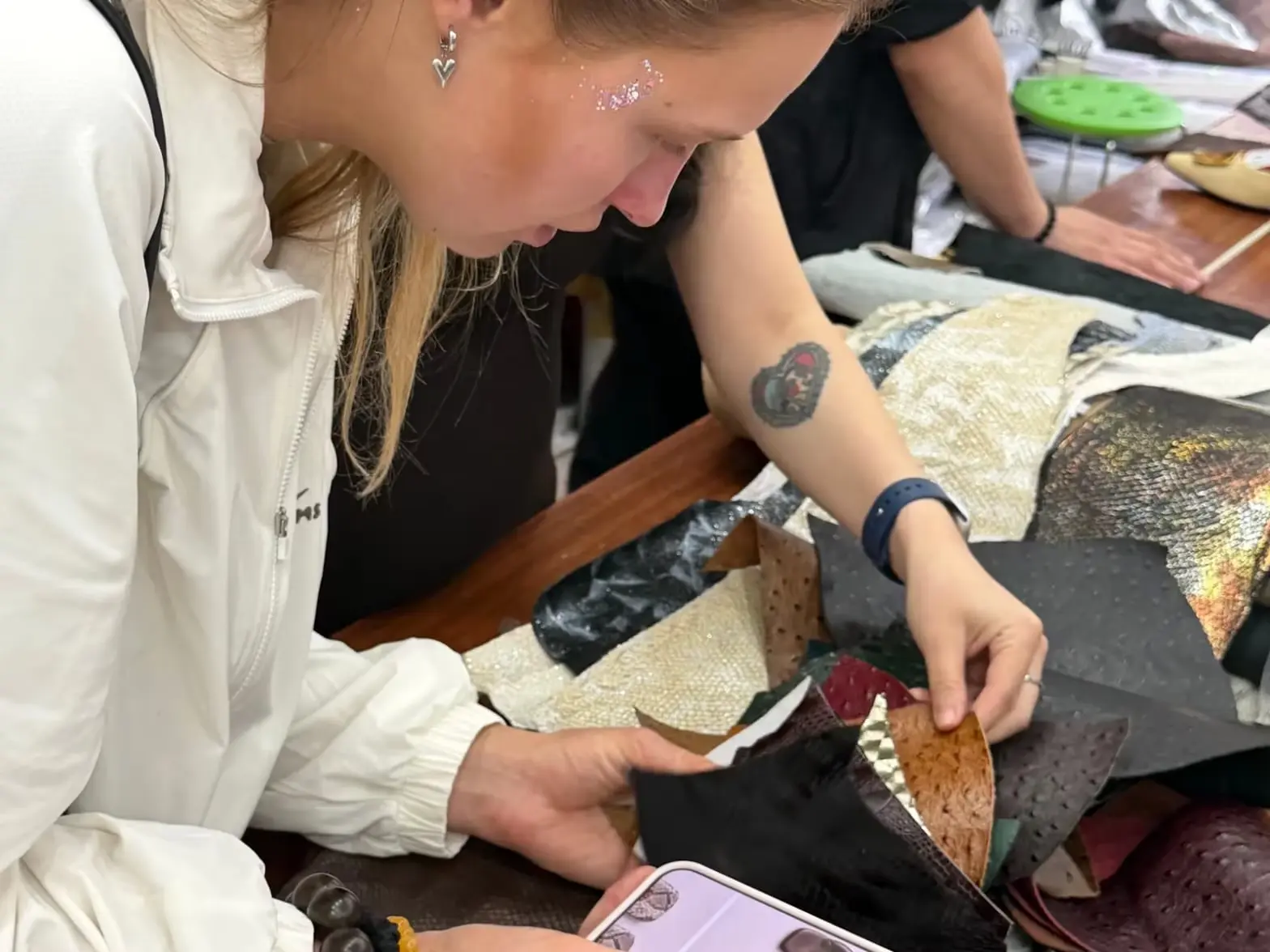 A designer bent over a pile of leathers, turning them over one by one, with the shoe design shown on a phone in her hand