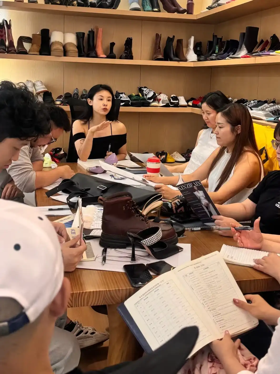 People seated around a long table in a development room with sketches, sample shoes, bags and a product book laid out, an open development ledger in the foreground