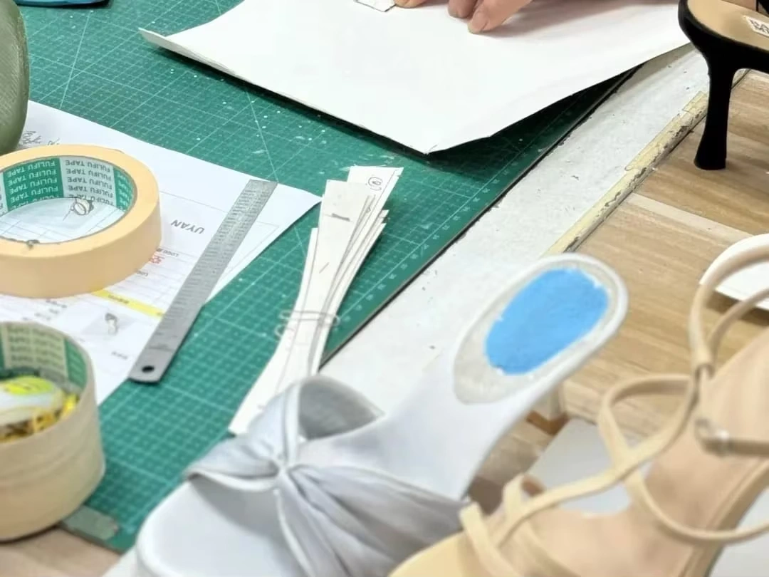 White paper patterns, pattern pieces, kraft tape, a steel ruler and a pen on a green cutting mat, with finished shoes at the edge