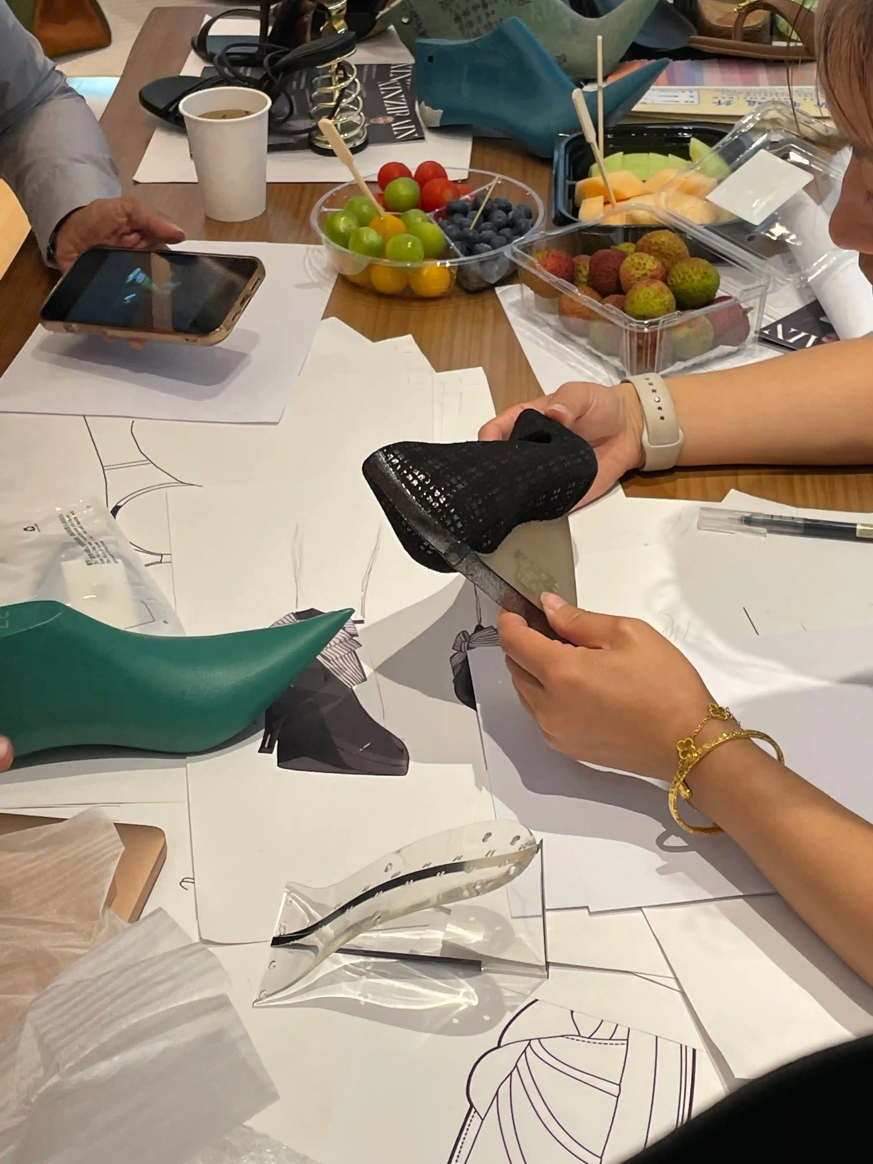 Two hands holding a black sequinned sample upper above a white shoe drawing for line-by-line comparison, green heel and sketches on the table