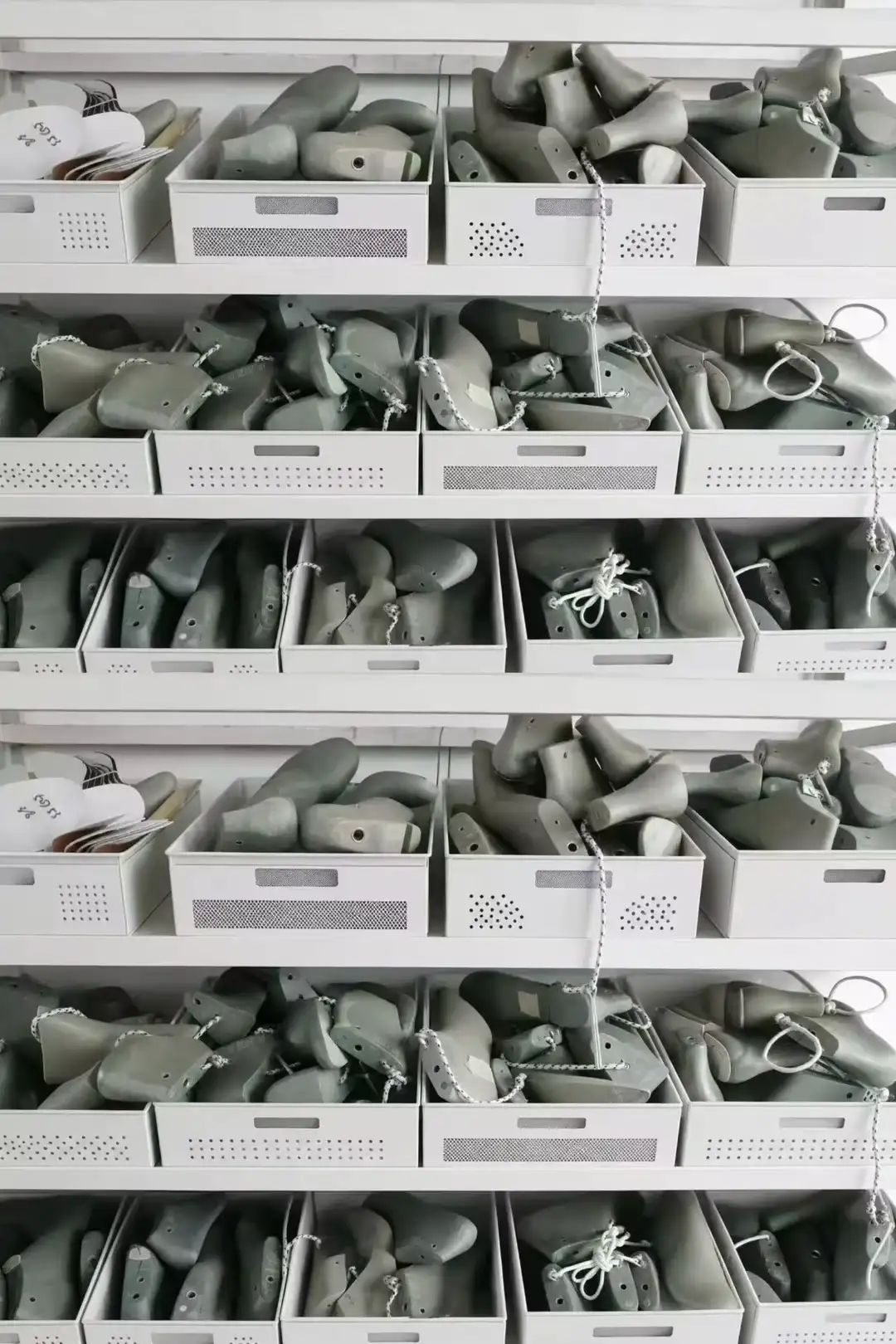 Rows of grey shoe lasts stored in white bins on shelving, sorted by style with string labels